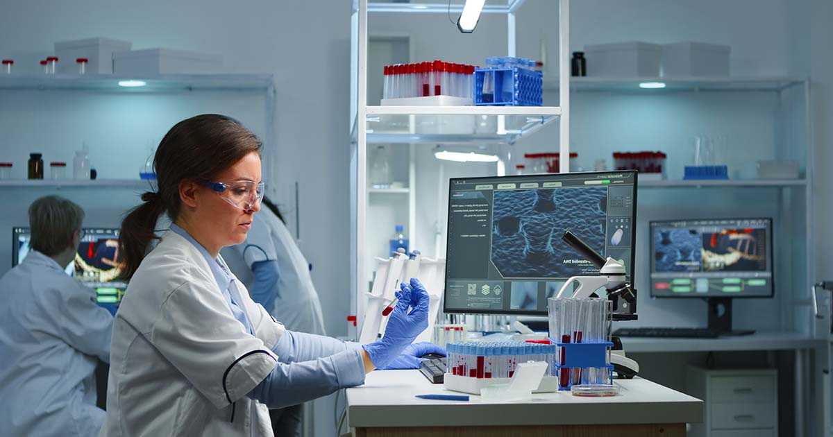 A woman in a lab, looking carefully at a vial in her hand. Two men are looking at a computer screen in the background